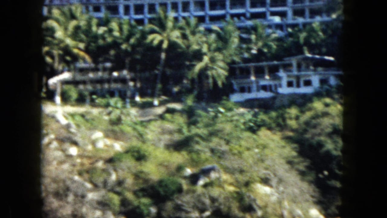 1962 Acapulco home-movie frame showing resort structures among palms on a steep shore above blue water.