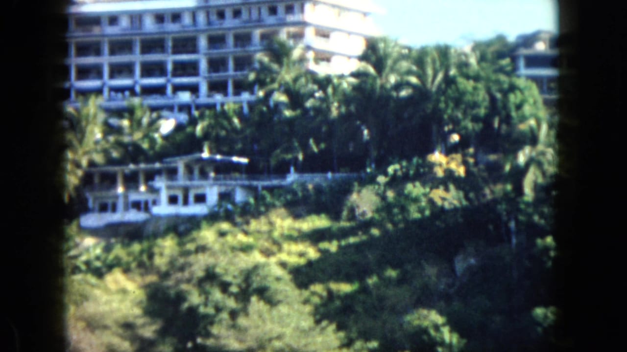 1962 Acapulco home-movie frame showing a white hillside hotel above palms, dense foliage, and a smaller light structure.