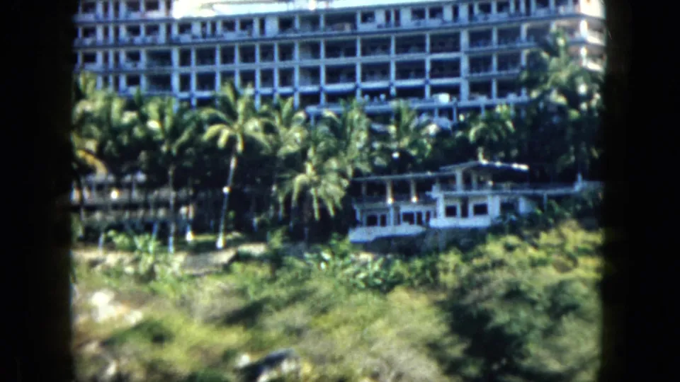 1962 Acapulco 8mm frame showing palms and thick greenery in front of a large white hotel on a hillside.