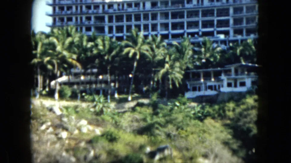 1962 Acapulco home-movie frame with a white hotel, tall palms, a small structure, and green hillside foliage.