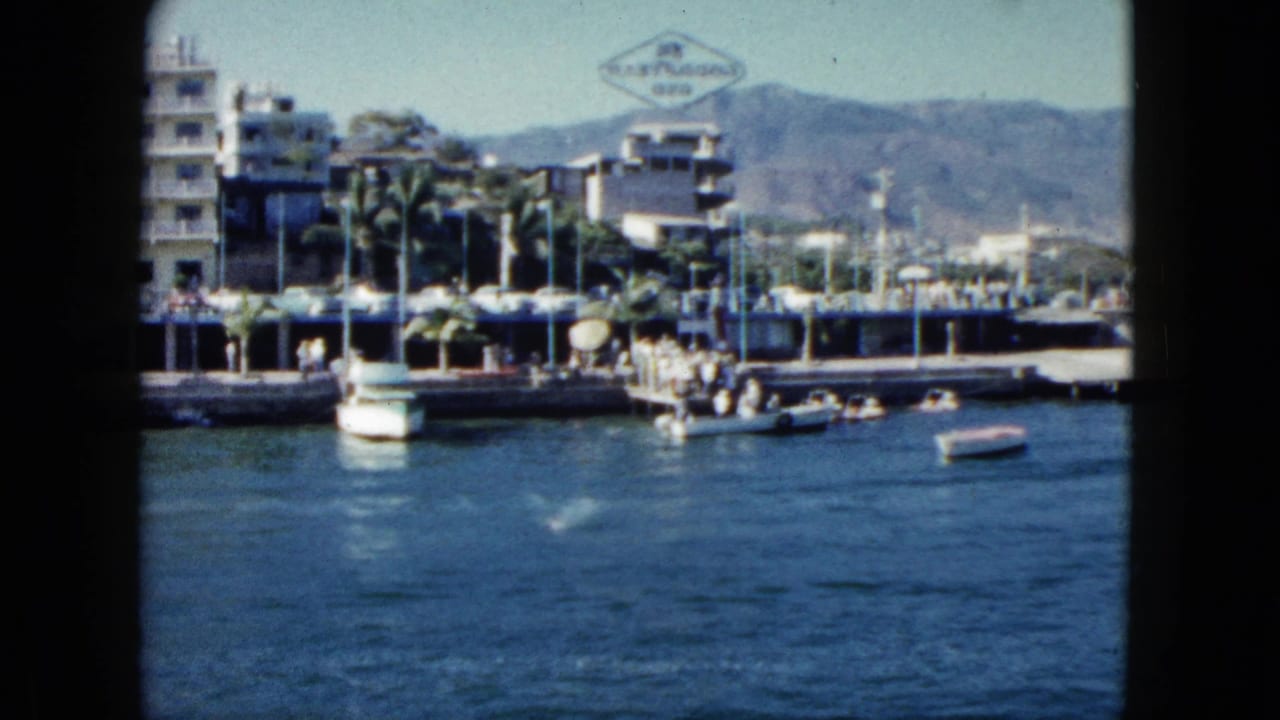 1962 Acapulco waterfront view with small boats near a pier, hotel buildings, palms, hills, and a visible hotel sign.