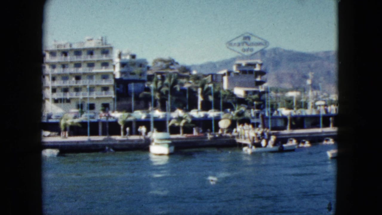 Acapulco harborfront in 1962 with boats on calm water, people near the pier, palms, hotel buildings, and hills beyond.