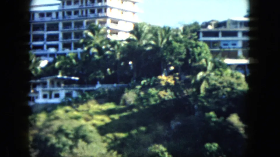 Acapulco shoreline in 1962, showing palms, hillside vegetation, and hotel structures viewed through soft handheld 8mm footage.