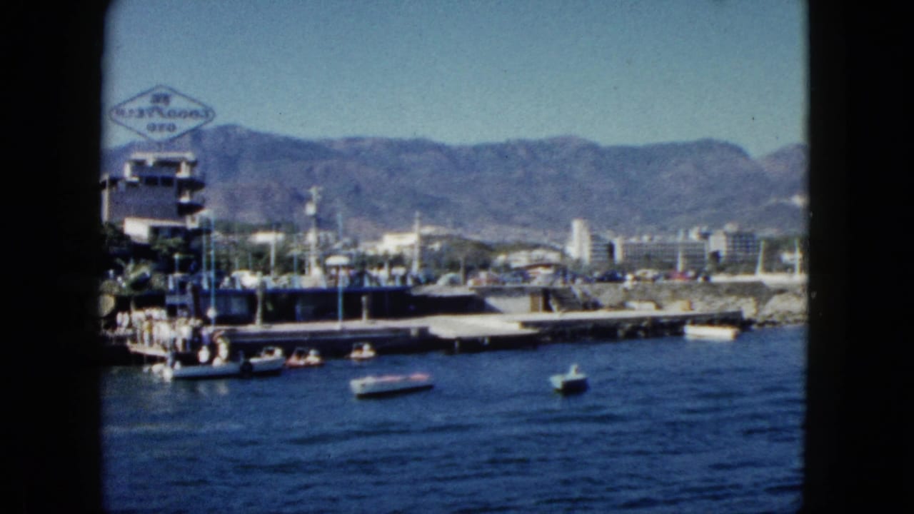 1962 Acapulco harbor viewed from the water: scattered motorboats, a commercial pier with a diamond-shaped sign, and arid sierra mountains under clear sky.
