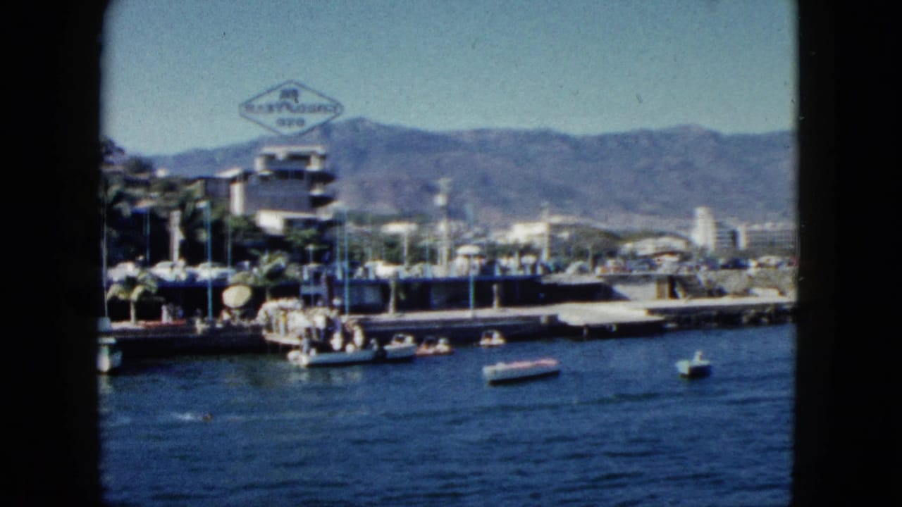1962 Acapulco waterfront: motorboats on calm bay, low-rise commercial buildings with a diamond-shaped sign, palm trees, and sierra mountains.