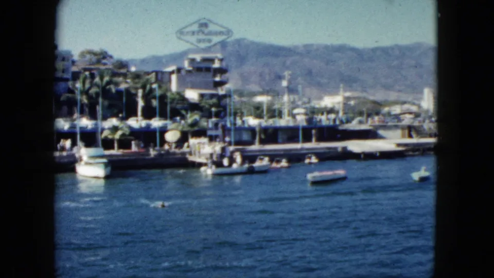 1962 Acapulco harbor: motorboats near a pier with a prominent diamond-shaped sign, palm trees, multi-story buildings, and figures visible on the waterfront.