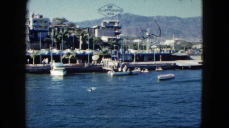 Final frame of a 1962 Acapulco harbor sequence: boats on calm water, a diamond-shaped sign above the pier, palm trees, and arid sierra mountains.