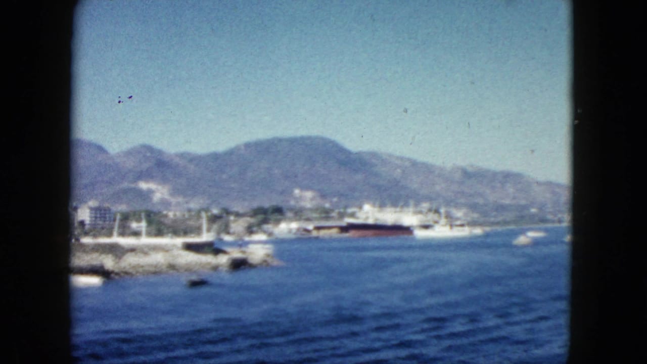 8mm view of what appears to be Acapulco marina circa 1962 — blue water, rocky breakwater, large ship at dock, white buildings, and rugged mountains under clear sky.