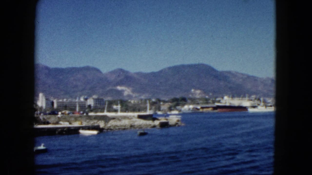 Harbor view through window frame with blue water, rocky shoreline, large docked ship, white buildings, and mountain backdrop.