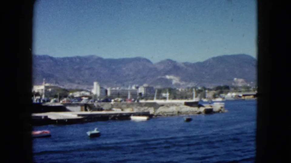 8mm view of Acapulco harbor circa 1962 — dock area with small boats and colorful objects near shore, white waterfront buildings, mountain range, and clear sky.