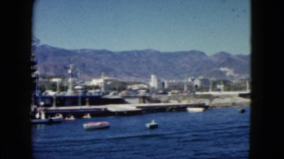 Acapulco marina circa 1962 on 8mm — pier structure in foreground, small motorboats on the bay, small figures near dock, white waterfront buildings, and rugged mountains.