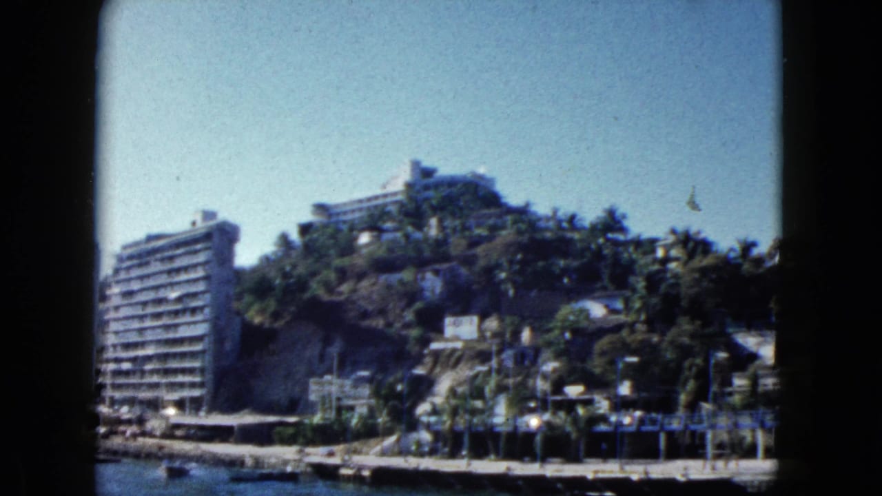 Large multi-story hotel on a tropical palm-covered hillside above a harbor waterfront, 8mm film.