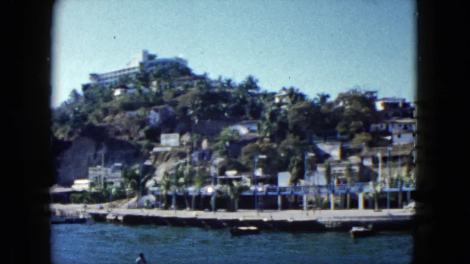 Acapulco, Mexico, 1962: wider hillside framing with modernist hotel, dense palms, and harbor dock below, on 8mm color film.