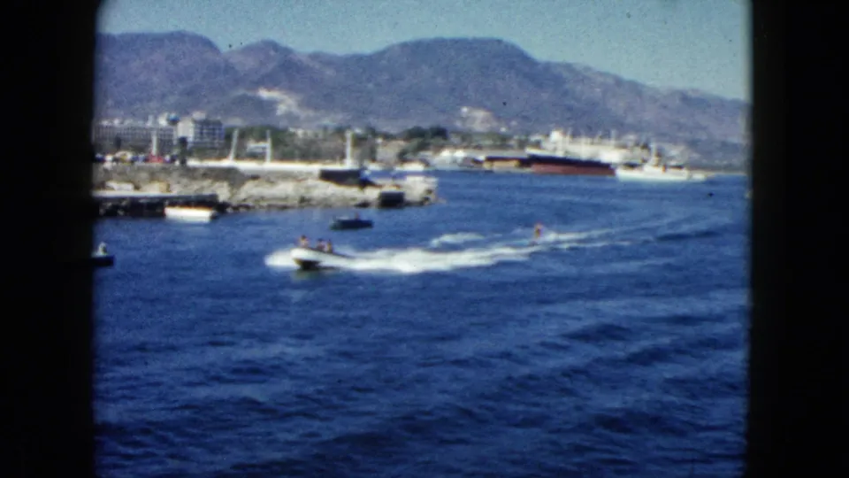 Acapulco Bay, 1962: speedboat crossing blue harbor water with a large ship at dock and rugged mountains in the background.