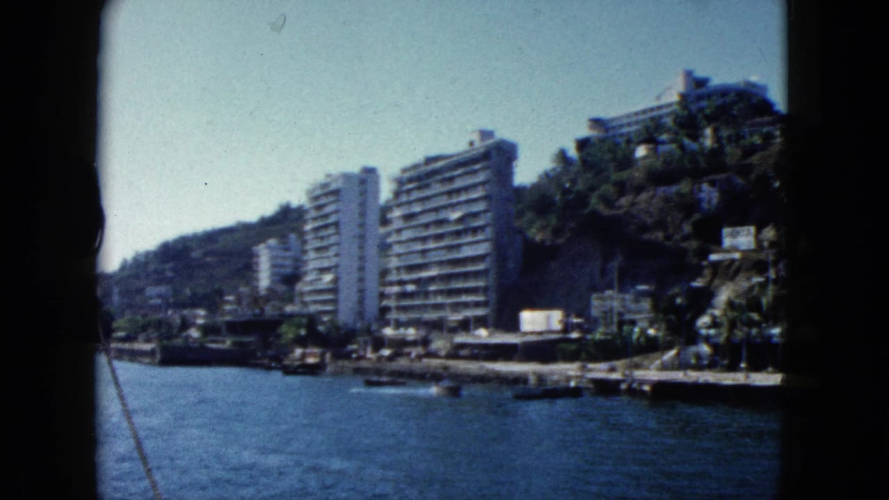 Continuing pan from water showing resort hotels on a steep green hillside above Acapulco Bay, open blue water in foreground.