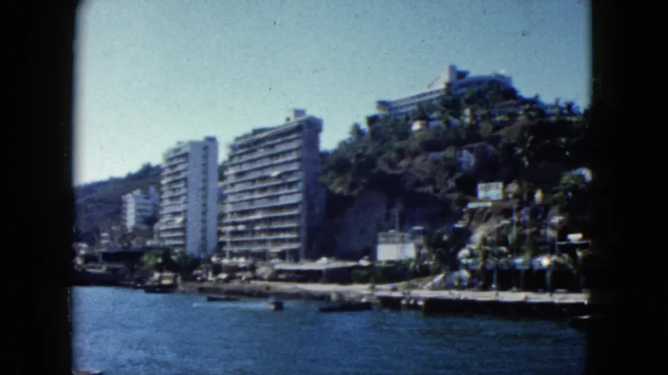 Boat-level pan showing a palm-lined waterfront promenade and multi-story resort hotels on the Acapulco hillside, circa 1962, 8mm home movie.