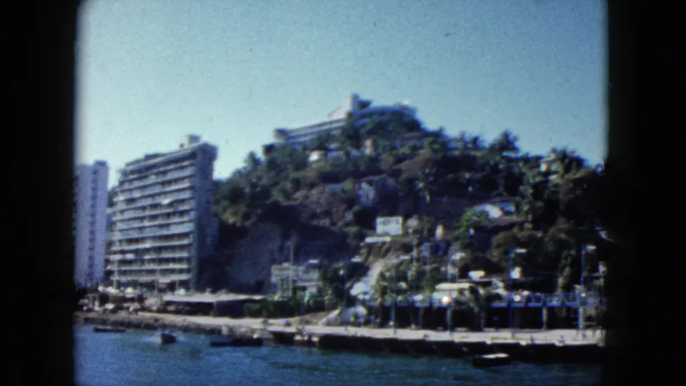 Closing pan across the Acapulco bay waterfront with resort hotels on a steep hillside and palm trees lining the shore, circa 1962, 8mm home movie.