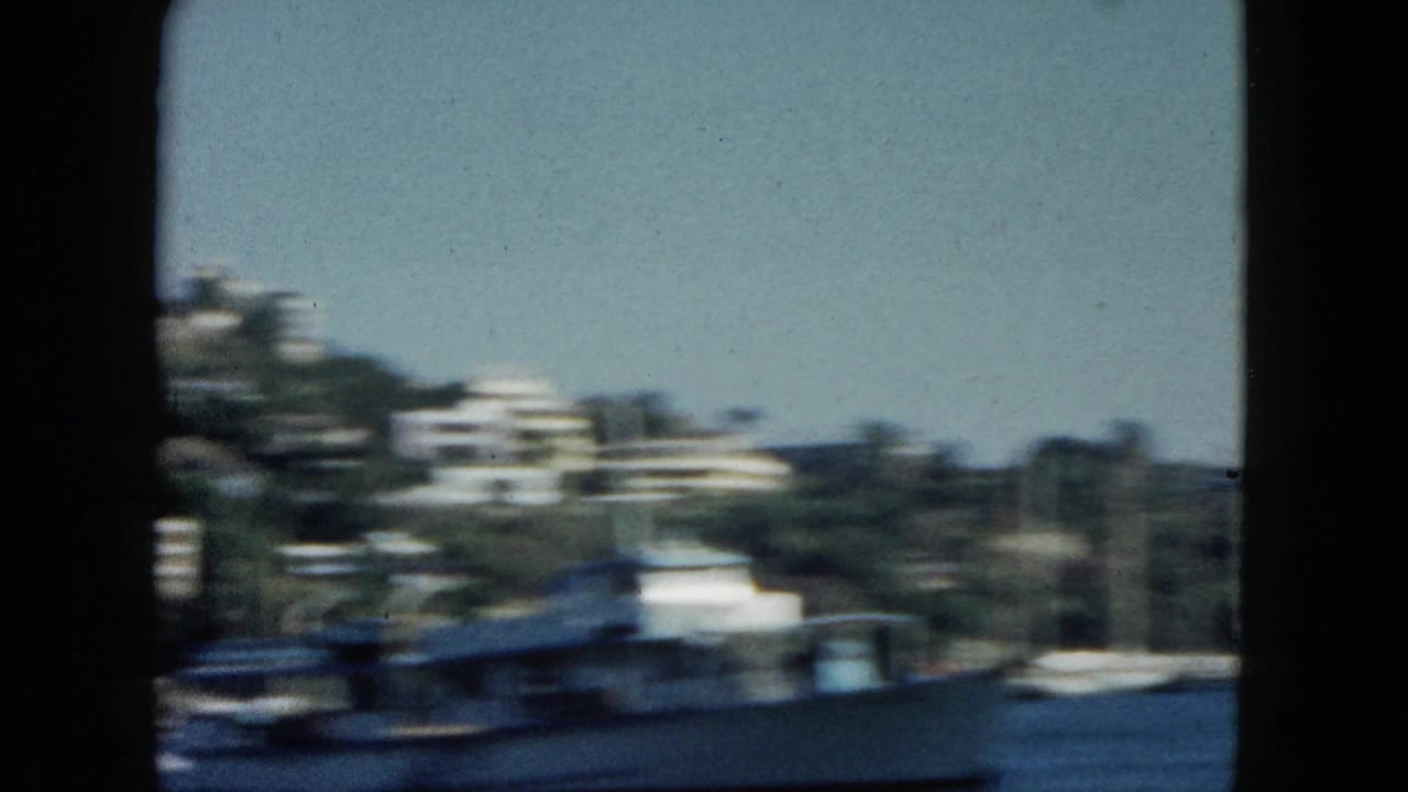 Large white boat blurred in foreground, white buildings on tropical hillside visible across the water.
