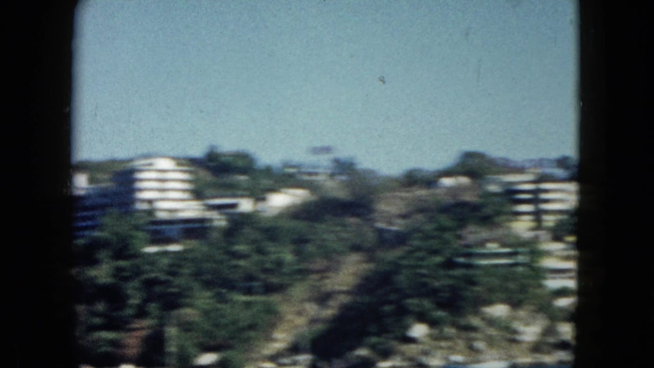 Multi-story white buildings with rounded balconies built into a steep tropical hillside, rocky slopes and palm trees surrounding them.