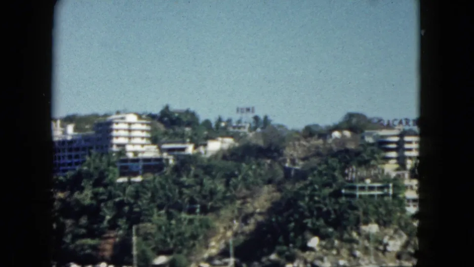 Wide pan across Acapulco bay waterfront, circa 1962 — large hotel building at left, palm-lined shore, rocky beach, and partially legible signage on seafront structures.