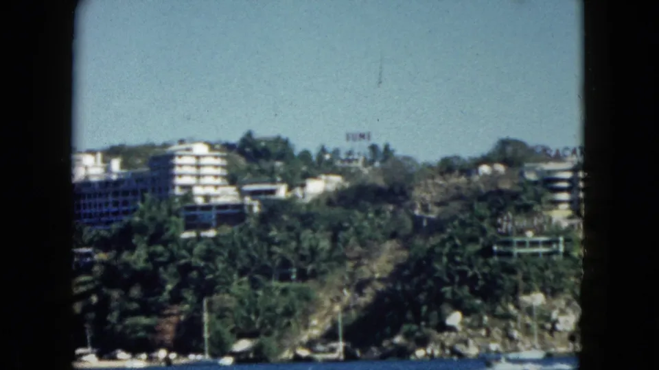 Late-pan frame across Acapulco bay, circa 1962 — multi-story hotel with balconies, palm trees, rocky coast, and illegible red signage on seafront buildings.