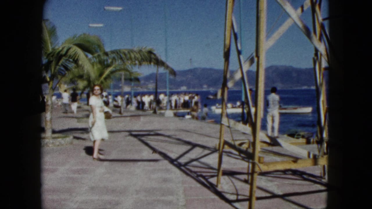 Woman in white dress strolling along a harbour pier in Acapulco, Mexico, circa 1962; palm trees, bystanders, and distant mountains frame the scene.