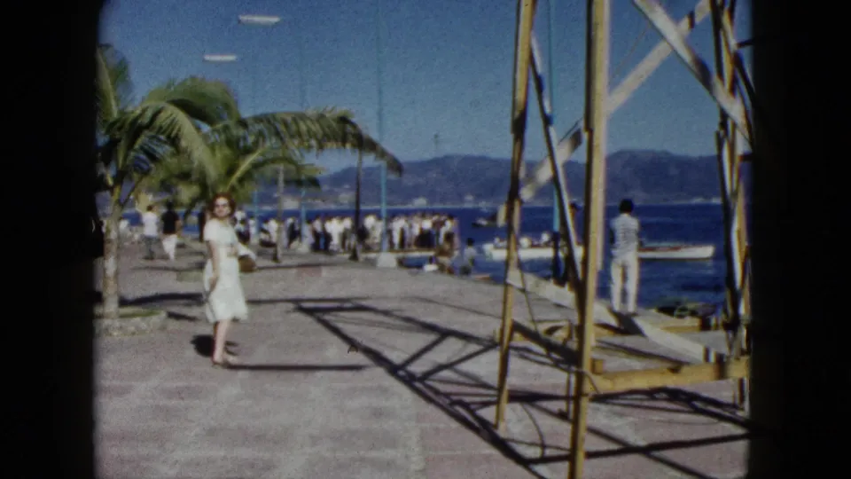 Woman in white dress strolling along a harbour pier in Acapulco, Mexico, circa 1962; palm trees, bystanders, and distant mountains frame the scene.