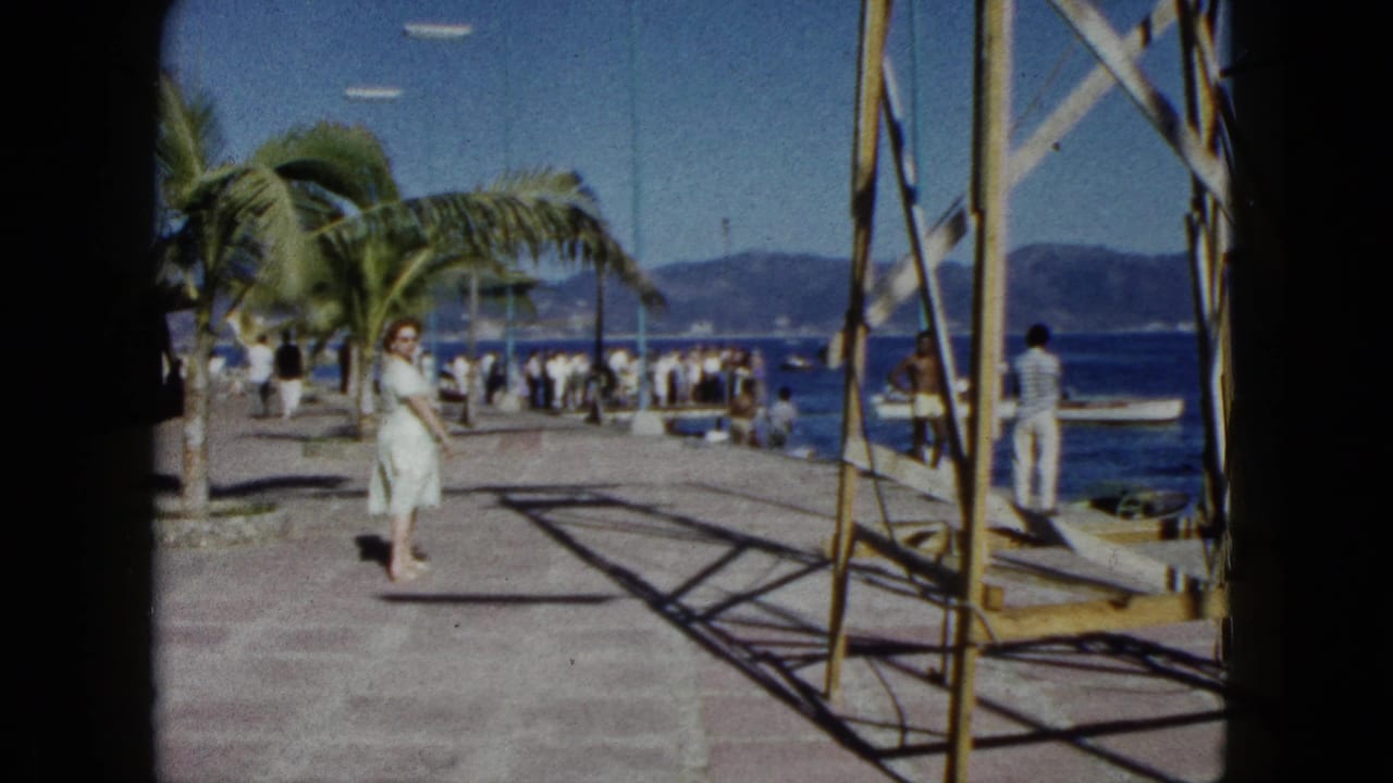 Woman in white dress continuing her walk along the Acapulco harbour pier, circa 1962; bystanders near the water and blue mountains visible behind her.