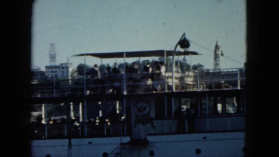 Passenger ship in Acapulco harbour, circa 1962; people on upper deck with a tower building and tree-covered hillside with coastal buildings in background.