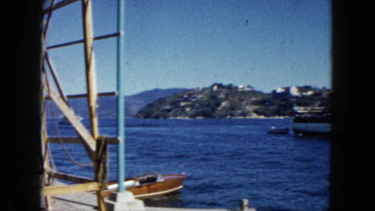 Wooden pier scaffolding frames a blue bay with a classic wooden motorboat moored at the dock; green hillside visible across the water.
