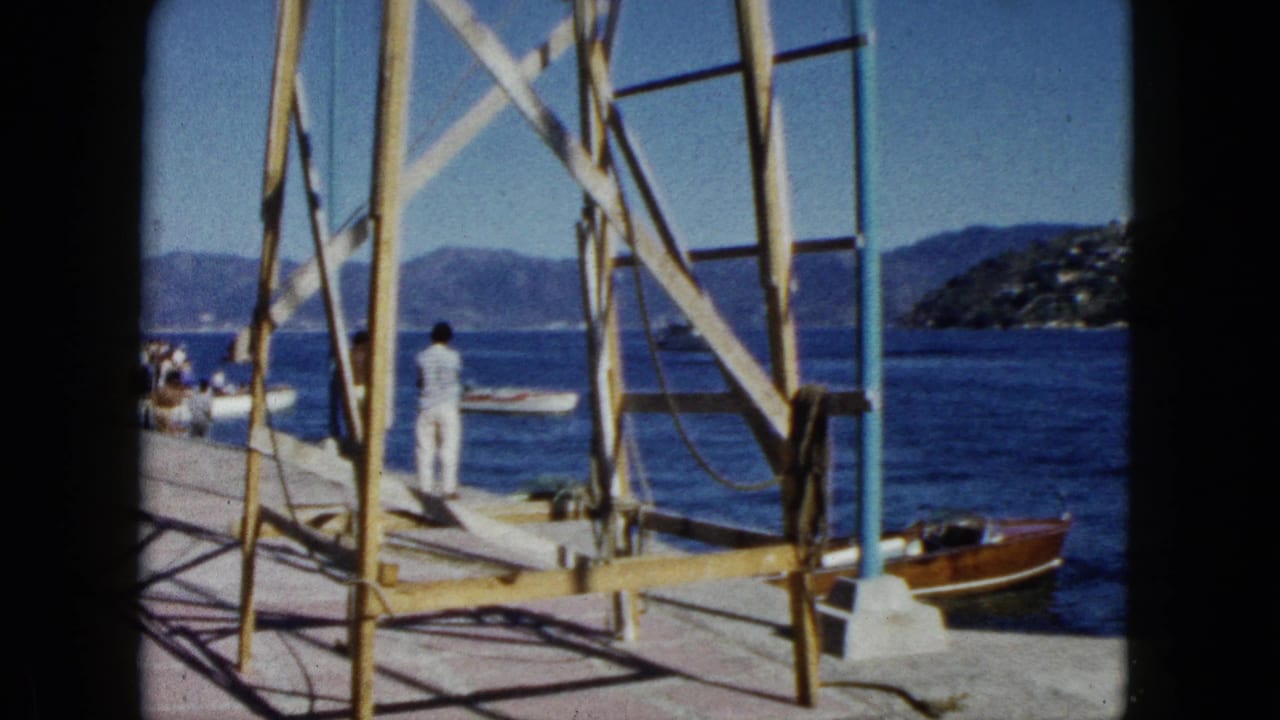 Figure in white stands on a paved waterfront pier beside wooden scaffolding; classic wooden motorboat moored to the right, distant hillside beyond.