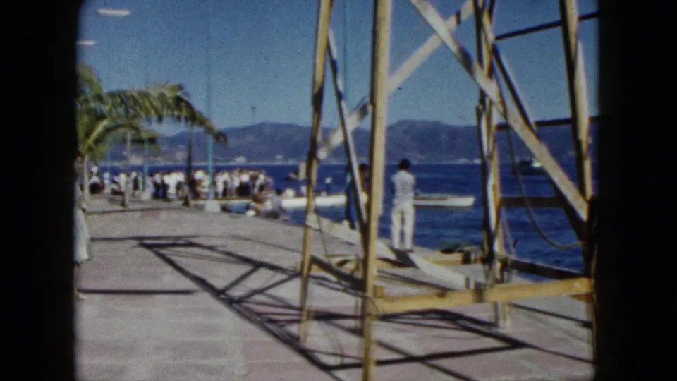 8mm home movie, Acapulco 1962: wooden dock scaffolding foreground, palm tree, crowd gathered on waterfront promenade, bay and green hillside.