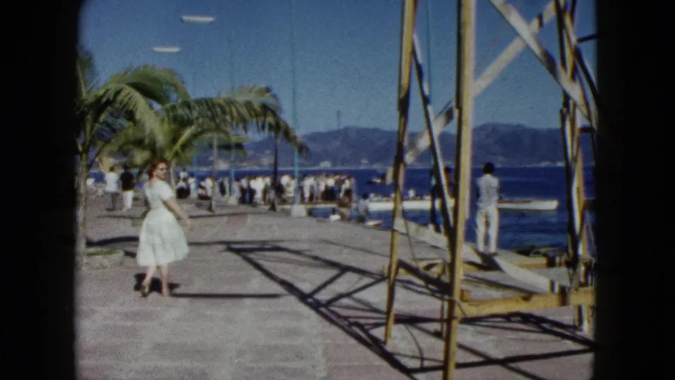 8mm home movie, Acapulco 1962: woman in white full-skirted dress mid-stride on waterfront pier, palm tree, scattered crowd, blue bay and hillside beyond.