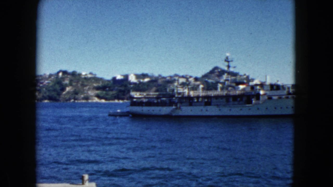 Large white multi-deck passenger vessel moored in calm bay, viewed through dark window framing, hilly coastline with white buildings behind.