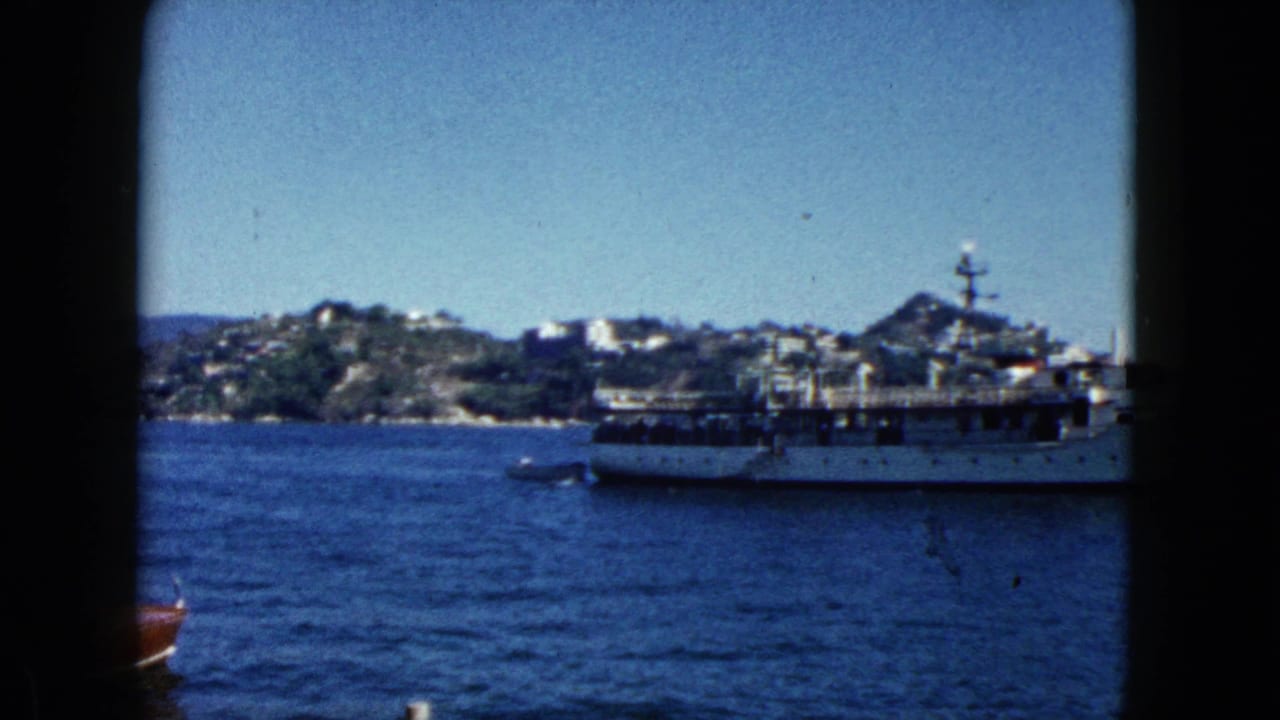 1962 Acapulco harbor in 8mm; large white passenger vessel at center with a small red-hulled boat entering the foreground, hilly coastline and white buildings behind.
