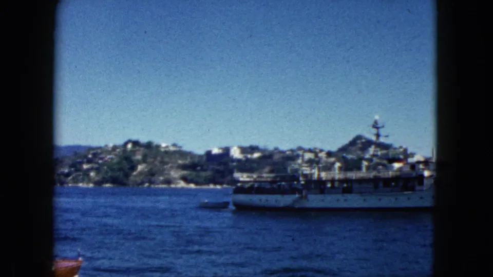 1962 Acapulco harbor, 8mm footage: large white ferry or passenger vessel in mid-ground and a wooden motorboat at frame bottom, rocky hillside with buildings under clear sky.