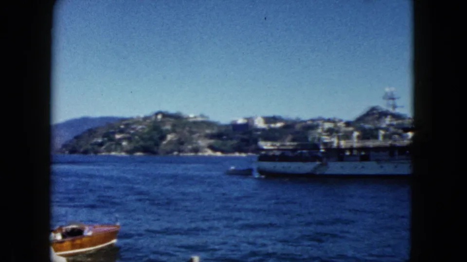 Final 8mm frame from 1962 Acapulco showing white vessel at right, wooden motorboat in foreground, and the rocky hillside with white buildings characteristic of Acapulco bay.