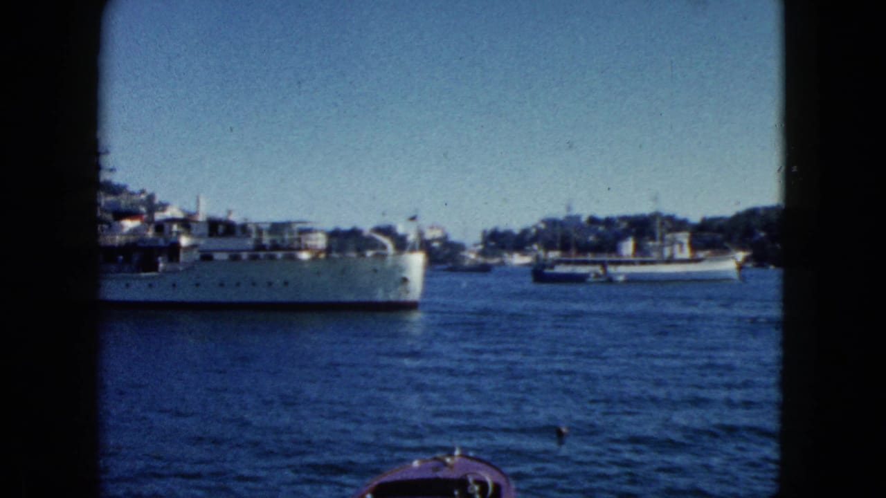 Large gray naval vessel in profile transiting Acapulco harbor, 1962; hillside with buildings and anchored smaller boats behind.
