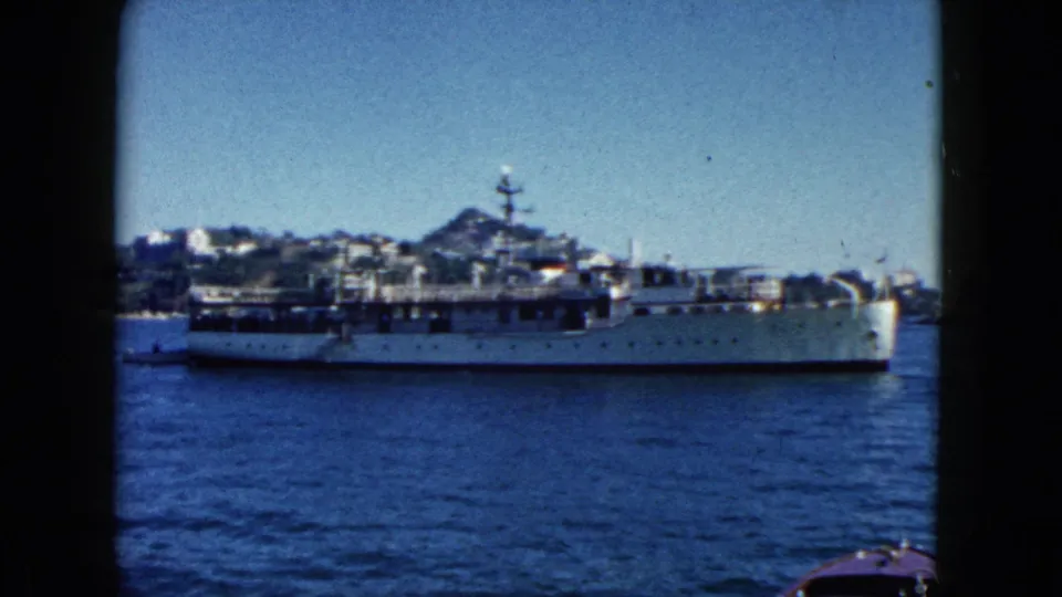 Gray naval vessel continuing passage through Acapulco harbor, 1962; rocky hillside with buildings increasingly prominent in background.