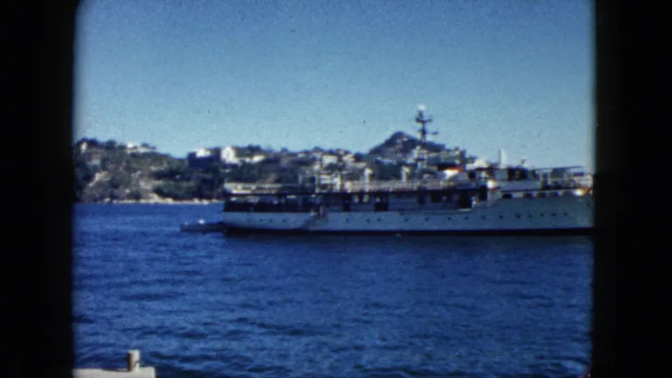 Naval vessel nearing exit of frame in Acapulco harbor, 1962; rocky hillside with clustered buildings fills background.