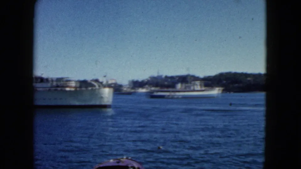 8mm travel footage c.1962, Acapulco harbour: large white passenger ship close in frame, tree-lined shore with low buildings, partial bow of a second vessel at lower edge.