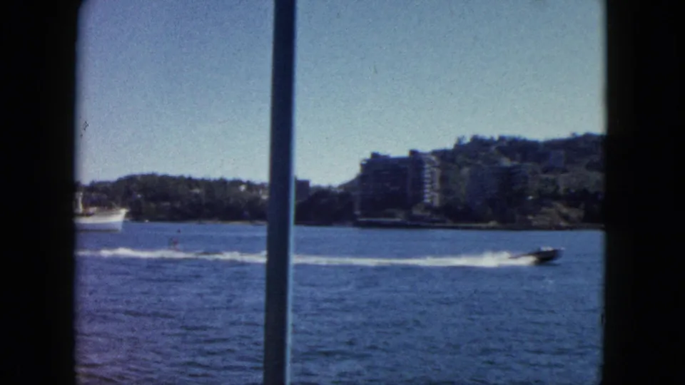 8mm home movie circa 1962, Acapulco bay: speedboat trailing white wake across blue water, early multi-storey buildings prominent on hillside.