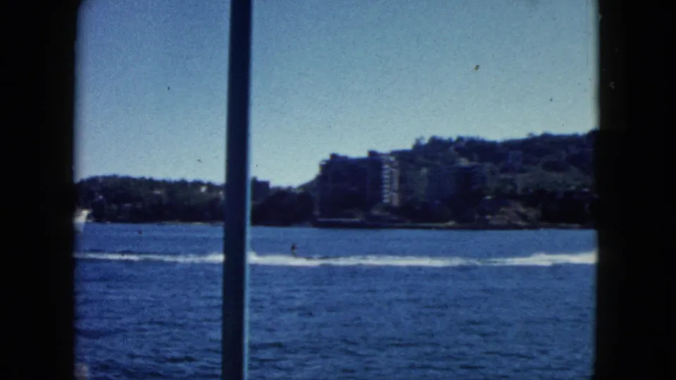 8mm home movie, Acapulco circa 1962: bay water with speedboat and wake in foreground; hillside shows early resort high-rise development under clear sky.