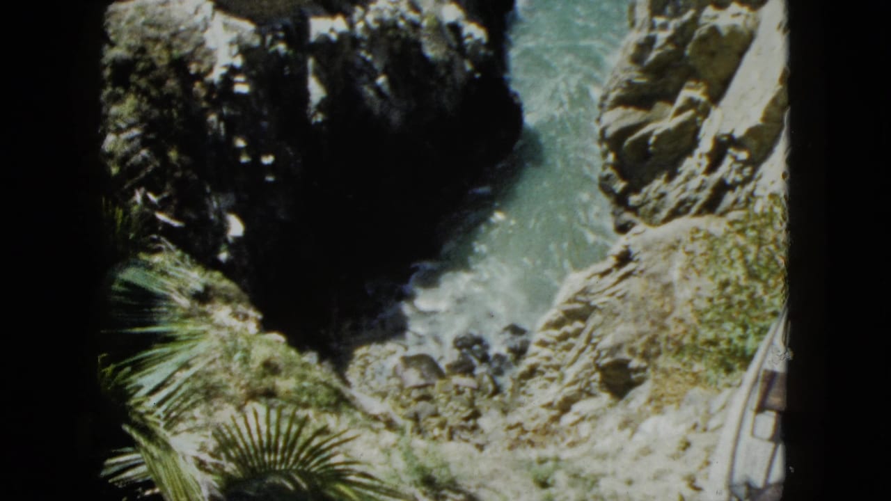 High vantage-point 8mm shot from an Acapulco cliff looking down into a rocky sea cove, turquoise water churning against jagged rocks, palm fronds at frame edge, circa 1962.