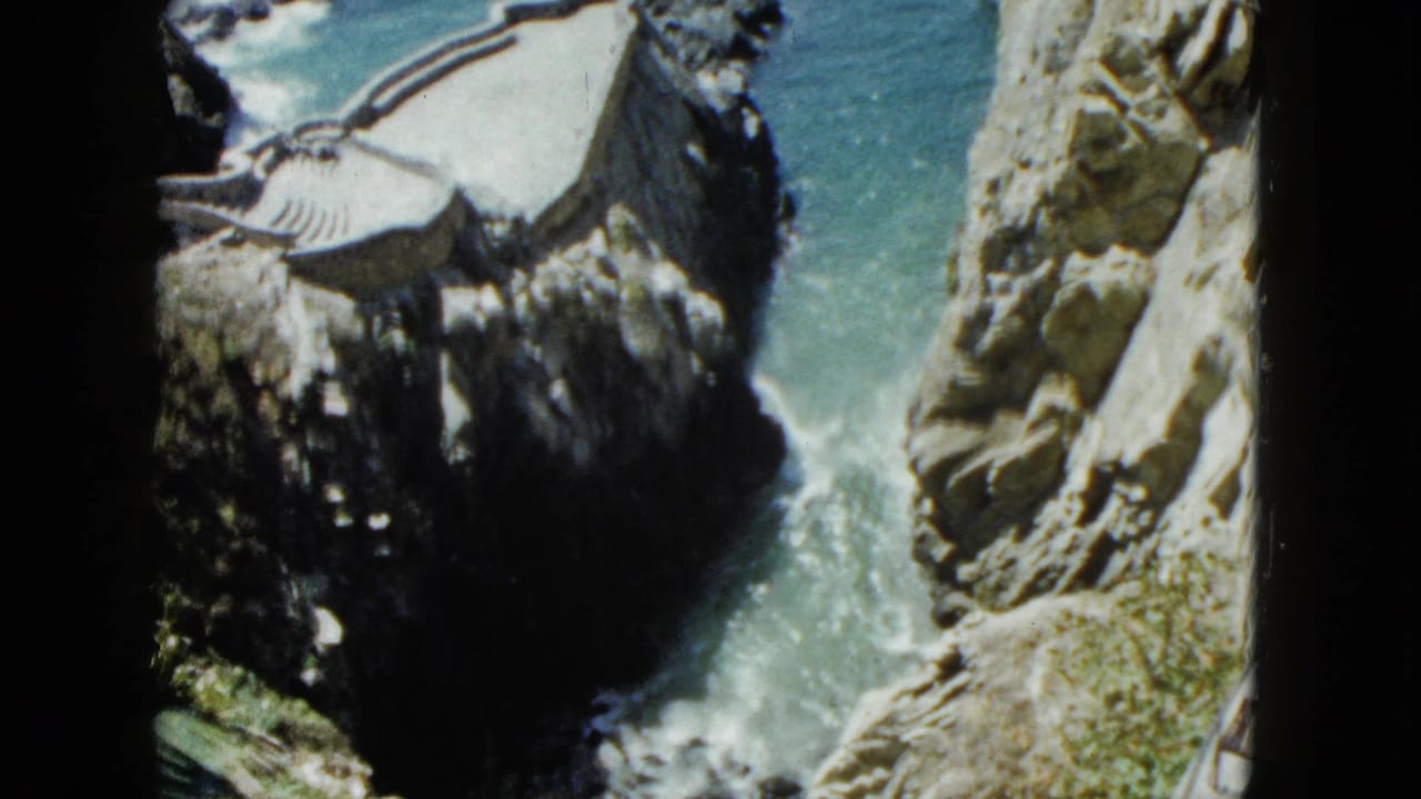 High-angle view down to a rocky cove with turquoise water; a small structure with a striped awning is visible at upper right on the cliff edge.