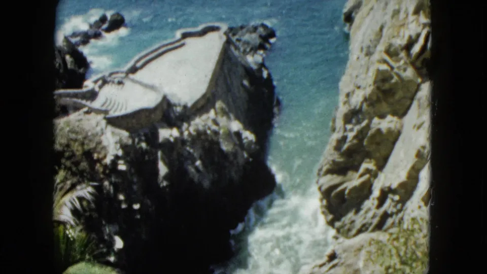 8mm footage circa 1962, Acapulco; a small platform or shelter with a striped awning is prominent at upper left above the narrow rocky cove and churning water below.