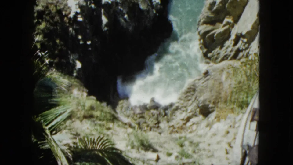 8mm home movie frame circa 1962, Acapulco; rocky cove with turquoise water, palm fronds filling the lower foreground, and a small cliffside structure with striped awning visible above.