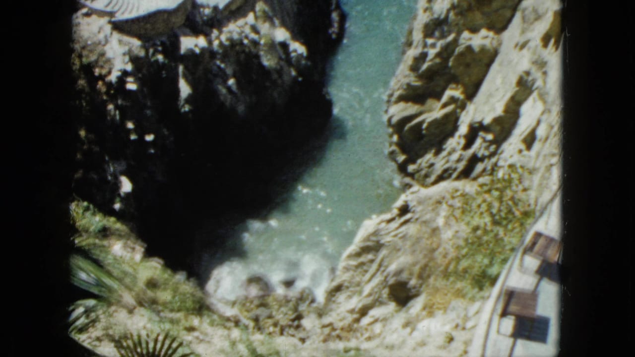 Downward view of rocky coastal cliffs with a narrow turquoise channel and white surf at the cliff base.