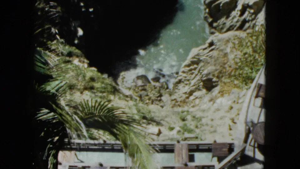 8mm frame, circa 1962 Acapulco: palm fronds frame the lower foreground while rocky cliffs and a narrow turquoise cove extend below, a wooden or metal railing visible at the cliff edge.