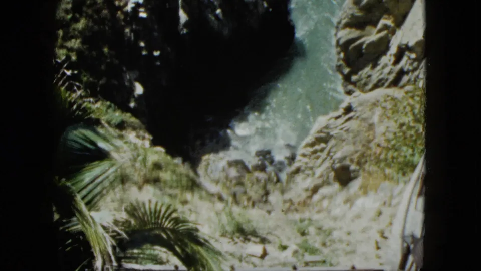 8mm frame, circa 1962 Acapulco: palm fronds lower right, rugged cliffs and turquoise sea channel below, railing of a clifftop structure visible, similar composition to prior frame.